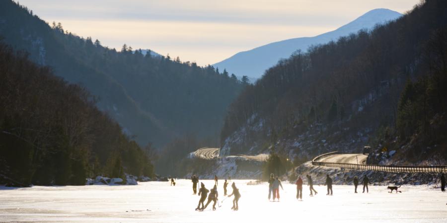 Group of people skating on a frozen lake with a country road and rolling mountains in the background